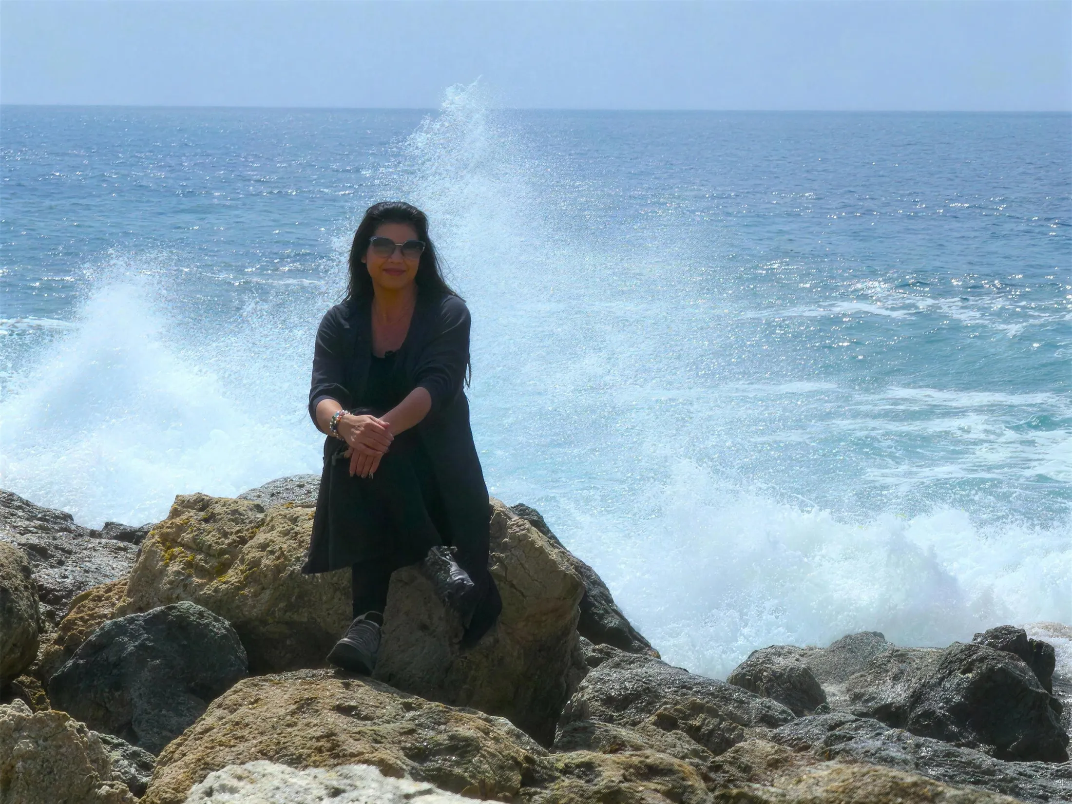 Sabine Kuhn sitzt gelassen auf Felsen am Meer, während hinter ihr eine mächtige Welle aufbricht